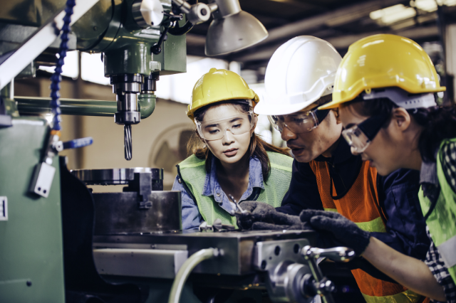 A group of people wearing hard hats, safety goggles, and construction vests looking at a piece of heavy machinery. 