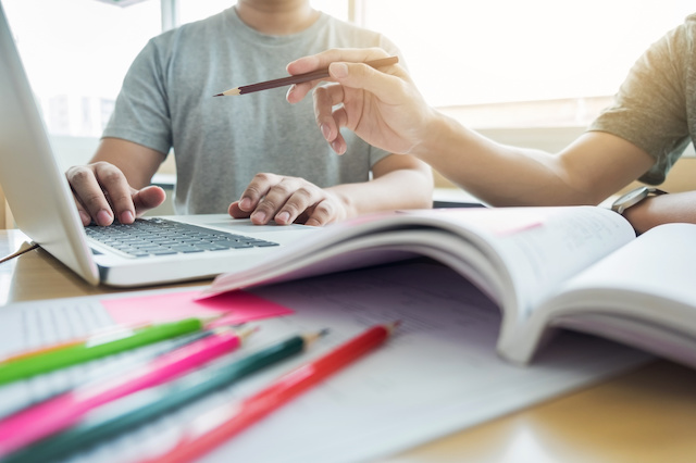 Two people sitting next to each other studying at a laptop. There is an open book, papers, and pencils laying on the table. 