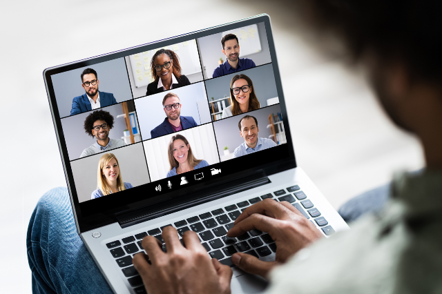 A person holding a laptop. The laptop screen has the faces of several people as if they're on a Zoom meeting. 