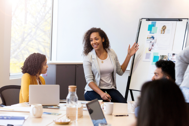 A woman gesturing toward a flip chart board with images and information on it. She is sitting at the head of a conference table with several people sitting around it.