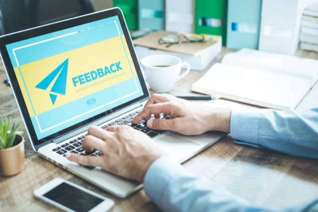 Close up of a person sitting at a work desk in front of a laptop. The laptop screen has a graphic on it of a paper airplane and the word feedback.