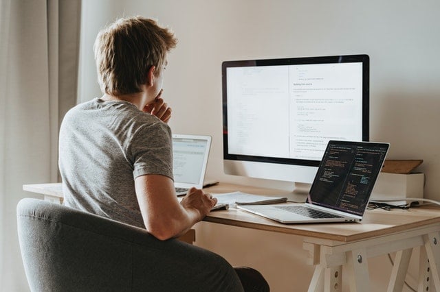 A person sitting at a desk with two laptops and a computer monitor on it.