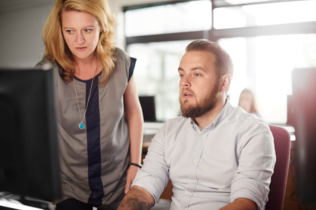 A man is sitting at a computer with a woman standing next to him. They're both looking at the computer screen.