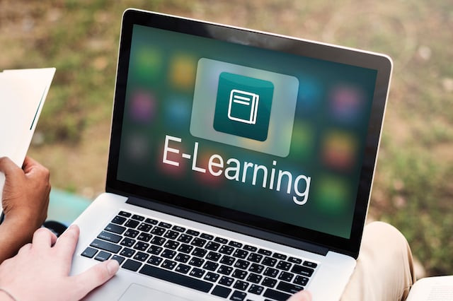 Close up of a person’s hands holding a laptop. The laptop’s screen features an icon of a book with the word “e-learning” written below. 