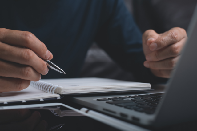 Close up of a person writing in a notebook with a pen while sitting in front of a laptop.