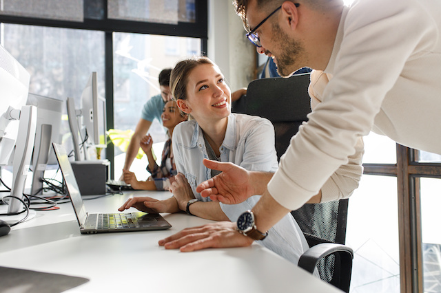 A group of adults are sitting at a long computer desk. The focal point of the photo is a woman sitting in front of her laptop. She is looking off to the right hand corner of the photo, where a man wearing glasses is looking down at her as if they are engaged in conversation.