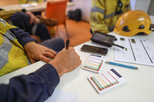 Close up of a person holding a pen while sitting at a table. There are notepads, a radio, a cell phone, and a yellow hard hat on the table.
